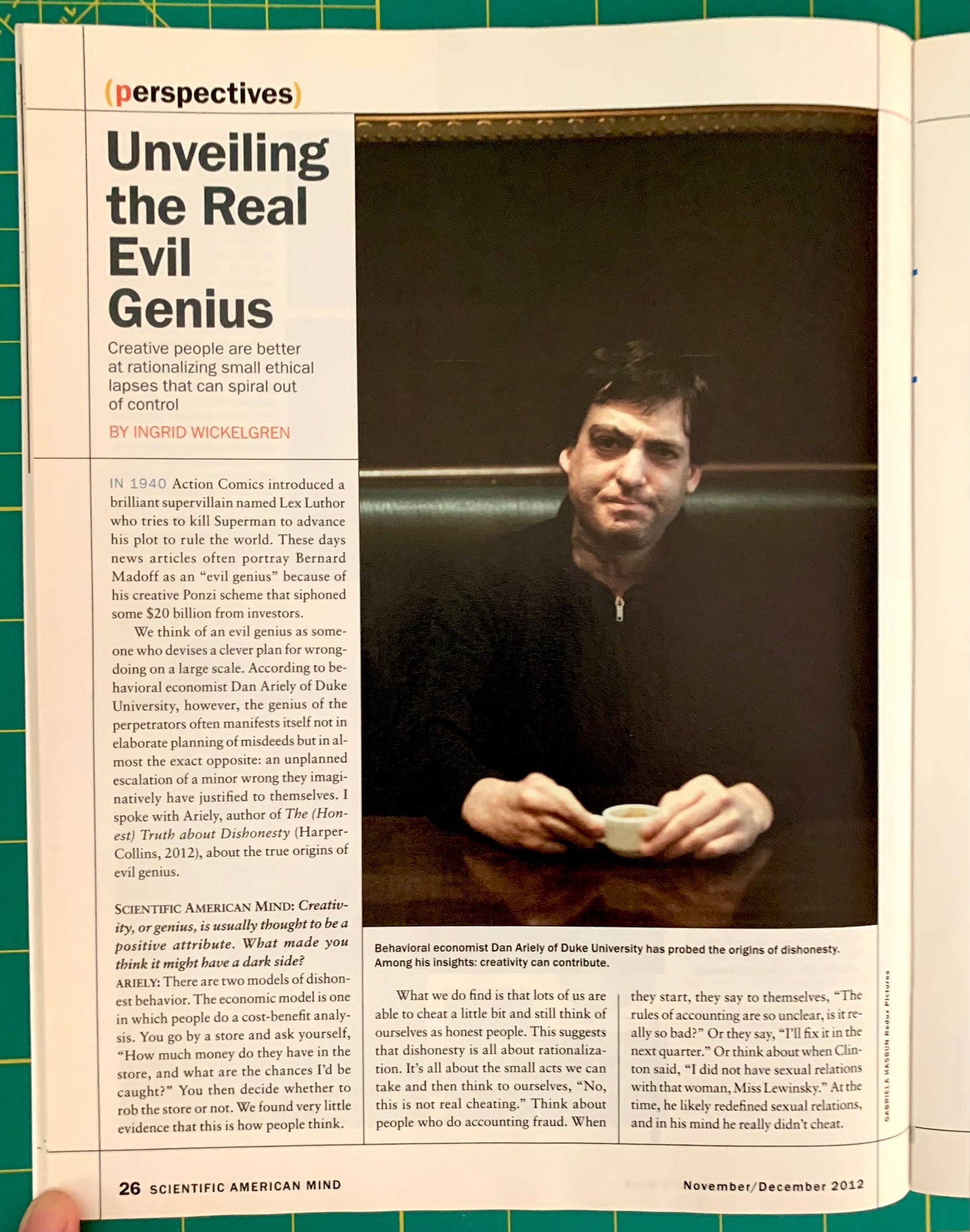A printed magazine or newspaper page spread shown open, featuring an article titled 'Unveiling the Real Evil Genius' with a large portrait photo of a man in a black suit and several columns of text. A hand is visible holding the page at the bottom.
[additional_image_link]