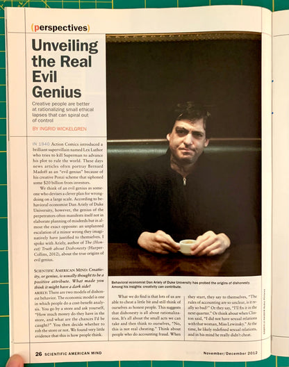 A printed magazine or newspaper page spread shown open, featuring an article titled 'Unveiling the Real Evil Genius' with a large portrait photo of a man in a black suit and several columns of text. A hand is visible holding the page at the bottom.
[additional_image_link]