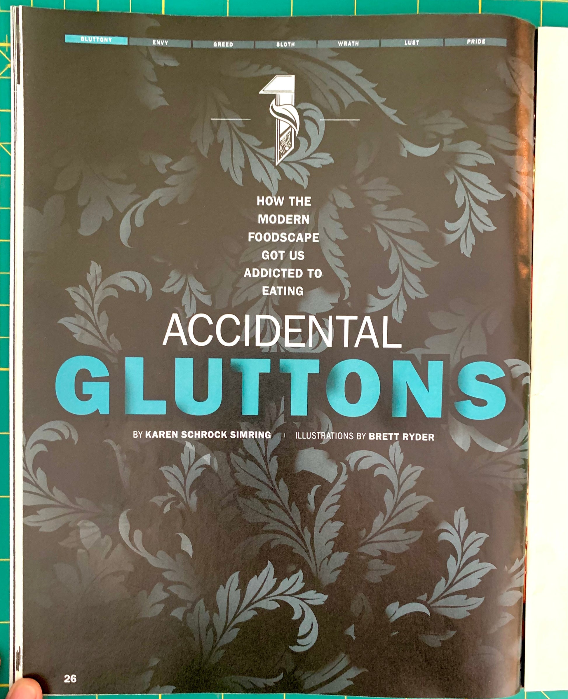 A magazine or book cover titled "Accidental Gluttons" with a dark brown background, pale gray ornamental leaf pattern, and large turquoise text. The cover is photographed flat against a green cutting mat background.
[additional_image_link]