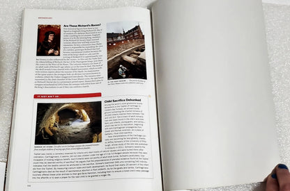 An open book showing printed pages with text, small photos, and a red headline, photographed on a light textured surface. A hand with dark nail polish is visible at the bottom right corner.
[additional_image_link]