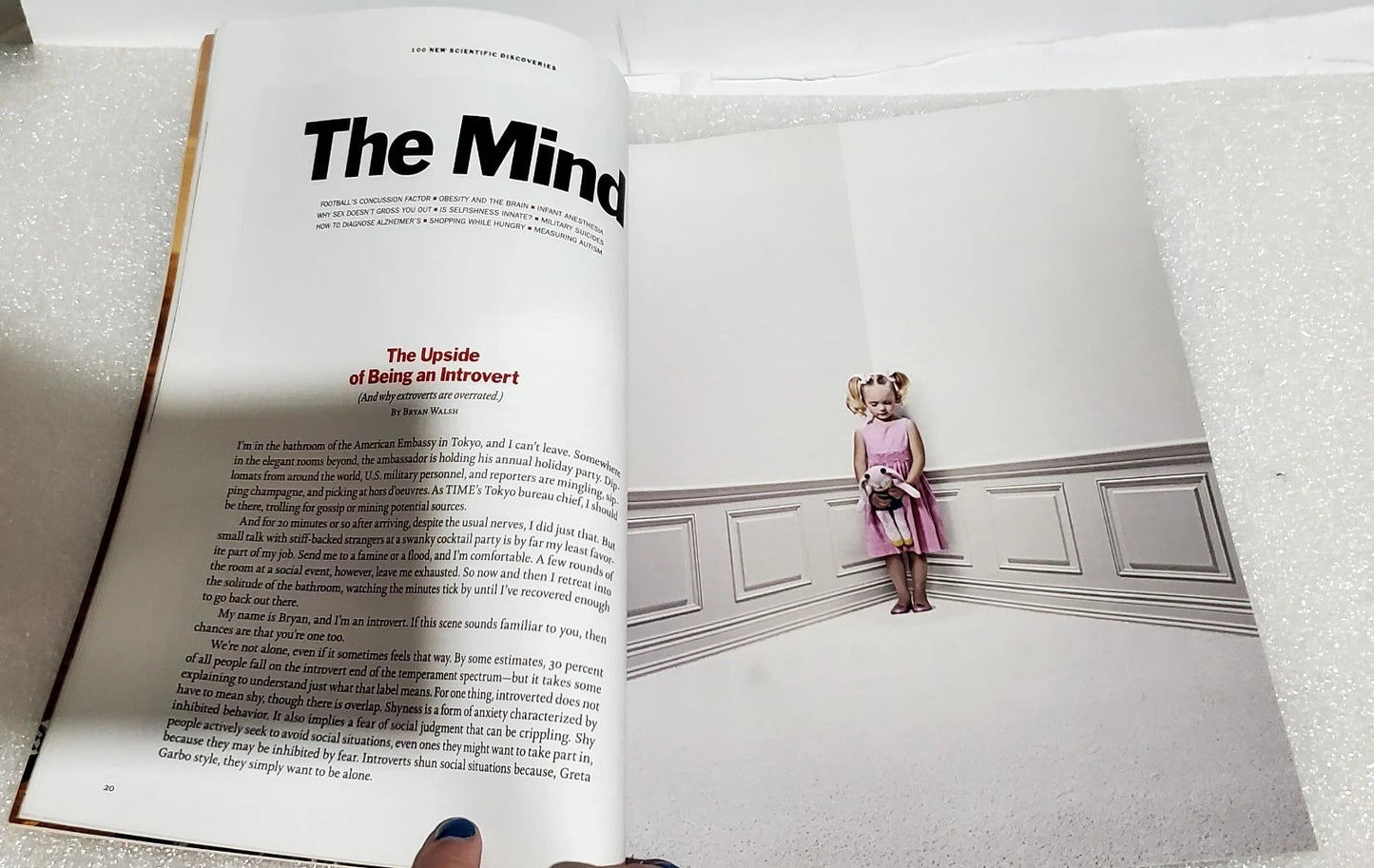 An open magazine or book spread showing printed text on the left page and a photograph on the right page of a young girl standing in a hallway. A hand is visible holding the open book at the bottom.
[additional_image_link]