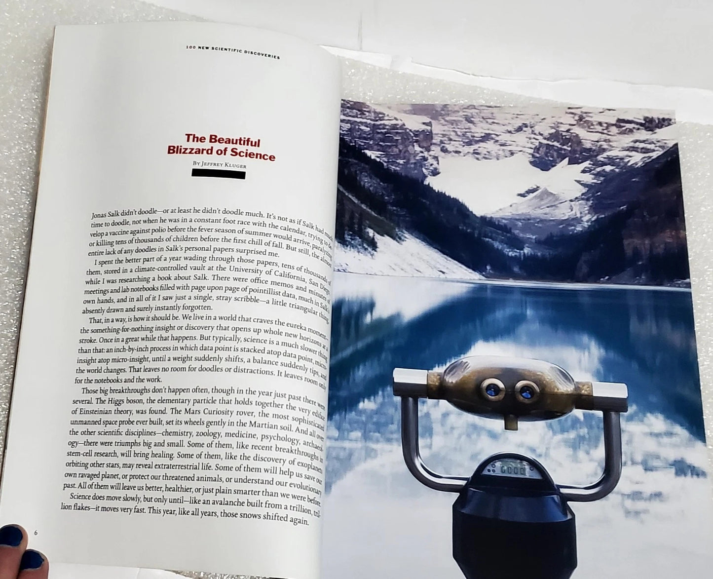 An open book or magazine spread showing a printed article titled 'The Beautiful Blizzard of Science' with text on the left page and a snowy mountain photograph on the right page. A small carved wooden figurine and a black object are placed in front of the page on a light surface.
[additional_image_link]