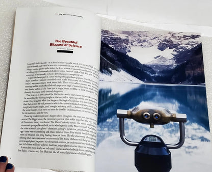 An open book or magazine spread showing a printed article titled 'The Beautiful Blizzard of Science' with text on the left page and a snowy mountain photograph on the right page. A small carved wooden figurine and a black object are placed in front of the page on a light surface.
[additional_image_link]