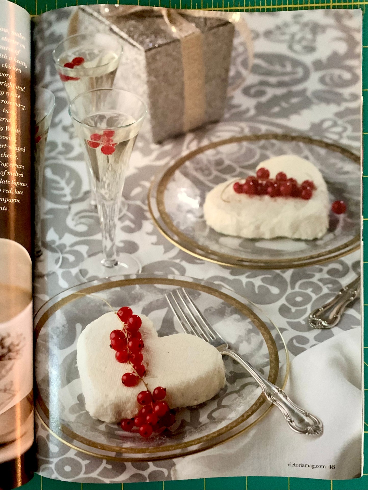 An open magazine or cookbook spread showing a styled dessert scene with two heart-shaped white cakes topped with red berries on gold-rimmed plates, champagne flutes, and a wrapped gift box on a patterned tablecloth.
[additional_image_link]