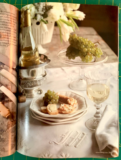 An open vintage cookbook or magazine spread featuring a styled table scene with a bottle of white wine, green grapes on a glass pedestal, plates of bread and cheese, and a filled wine glass, set against a green grid background.
[additional_image_link]