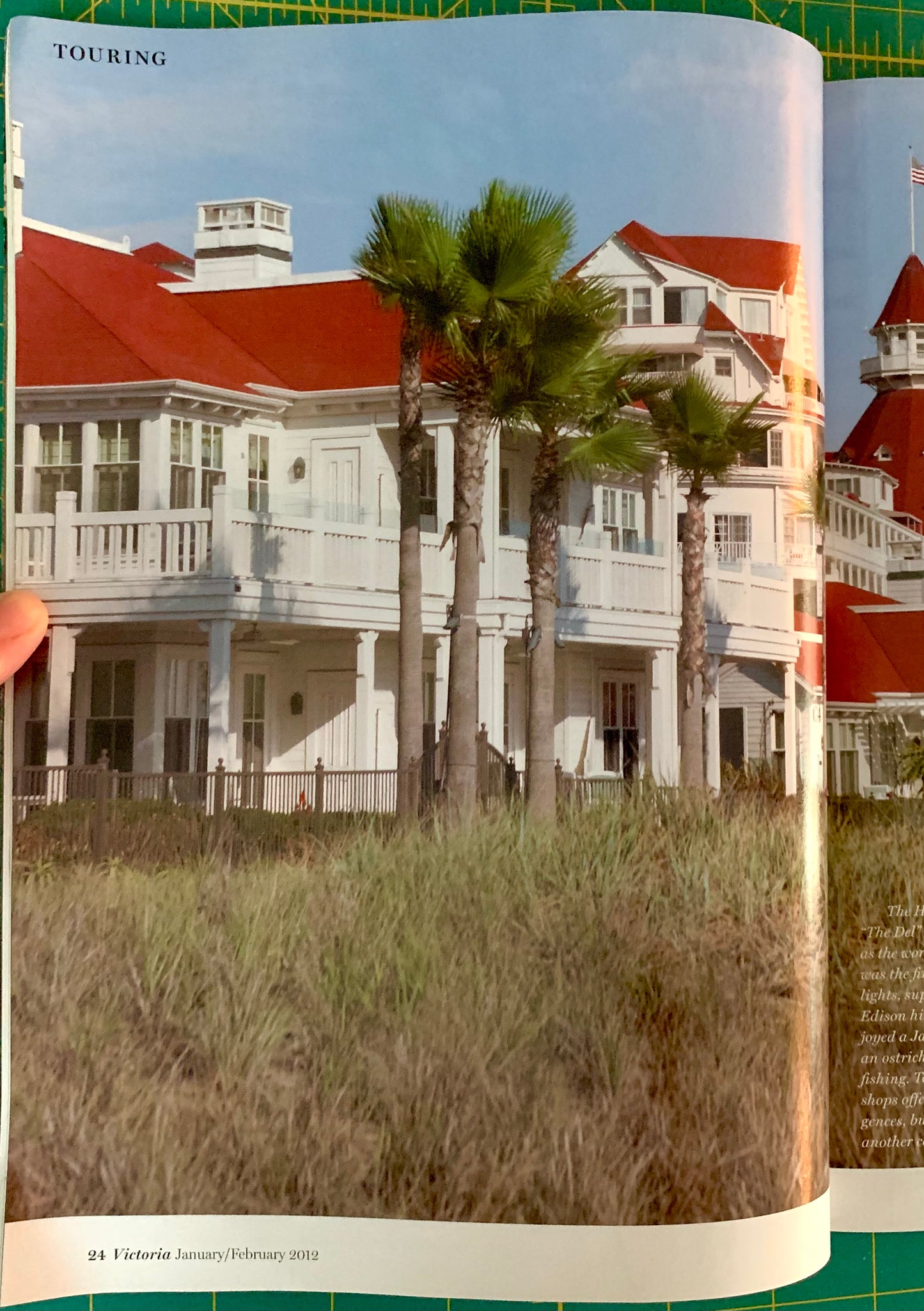 A printed page from a magazine or travel book showing a large white seaside mansion with red roofs and palm trees, photographed in situ on a green cutting mat with a finger visible at the left edge.
[additional_image_link]