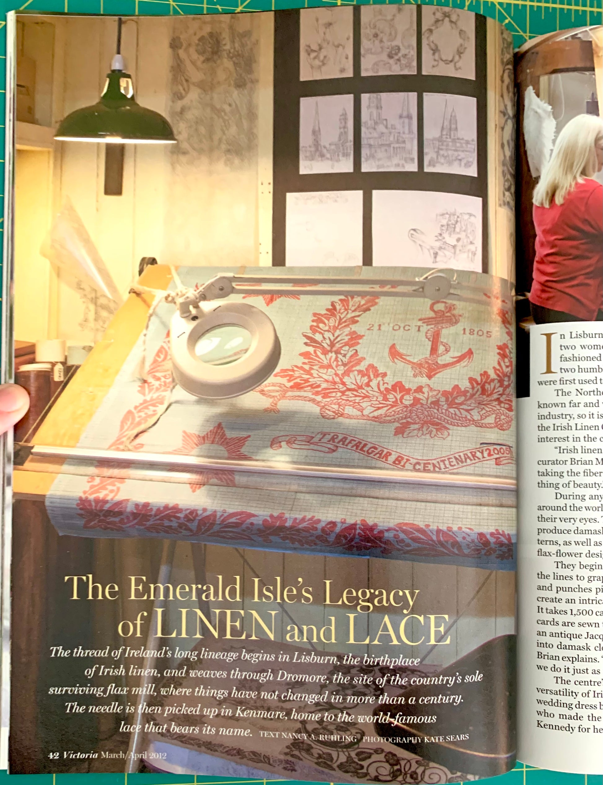 An open magazine or book spread showing an interior scene with a large sewing machine on a table, wallpaper, framed prints, and text headings including 'The Emerald Isle's Legacy of LINEN and LACE.' A hand is visible holding the left page edge.
[additional_image_link]