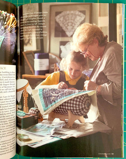 An open book showing a color photograph of an elderly woman and a child sewing a patterned quilt on a table, with printed text visible on both pages.
[additional_image_link]