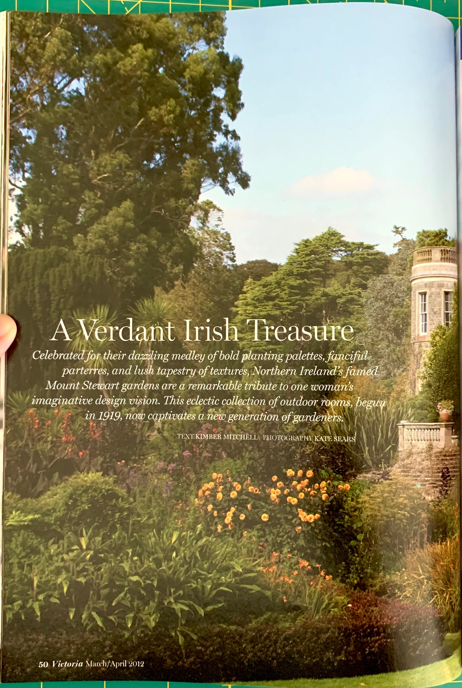 A printed page or book spread showing a lush garden scene with trees, shrubs, flowers, and a stone tower, overlaid with the headline 'A Verdant Irish Treasure' and several lines of descriptive text. A hand is visible holding the page at the left edge.
[additional_image_link]