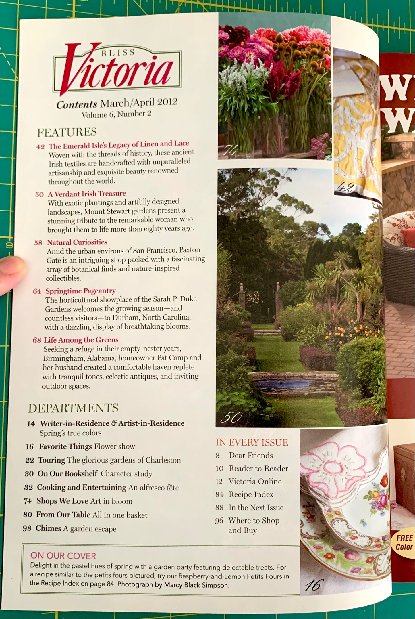 A hand holding a printed magazine or book page featuring the Victoria recipe collection, with a floral kitchen scene printed on the right page and visible text sections like Features and Departments.
[additional_image_link]