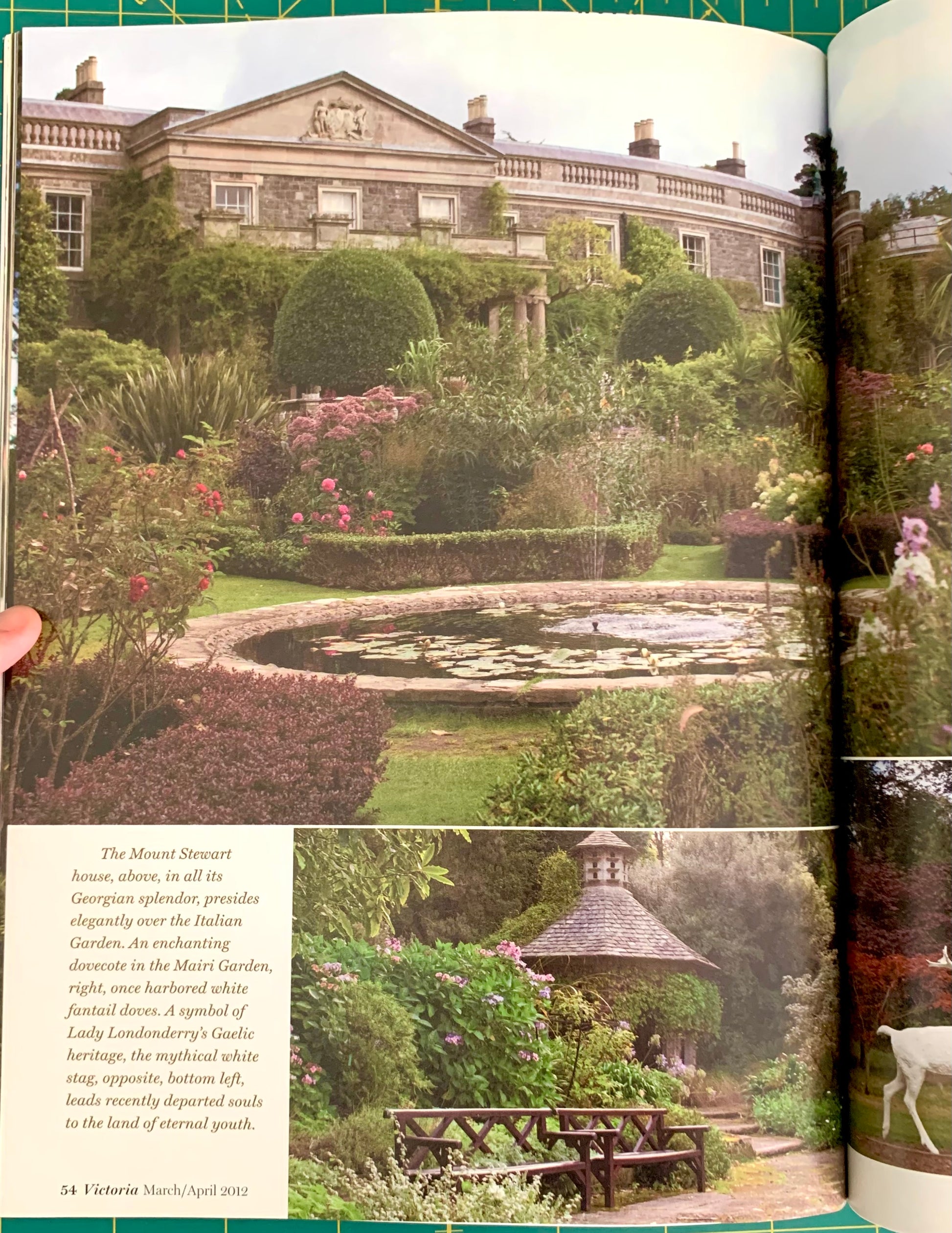 An open book showing a printed page with a large photograph of a formal garden and stone mansion, plus smaller inset photos and a block of text. A hand is holding the left edge of the book.
[additional_image_link]