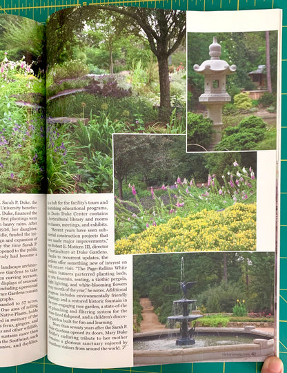 An open book showing a garden landscape photograph with text columns and inset photos of a stone lantern and flowering plants, photographed on a green cutting mat with a finger visible at the bottom right.
[additional_image_link]