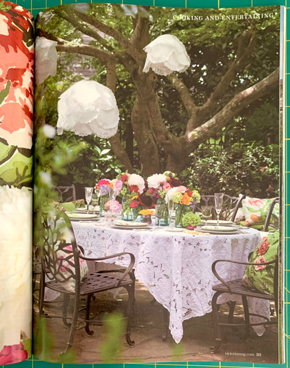 A printed page from a magazine or book showing an outdoor dining table set with a white lace tablecloth, metal chairs, and colorful flower arrangements under leafy trees. The left edge of the page shows a floral patterned fabric, and the page is photographed on a green grid surface.
[additional_image_link]