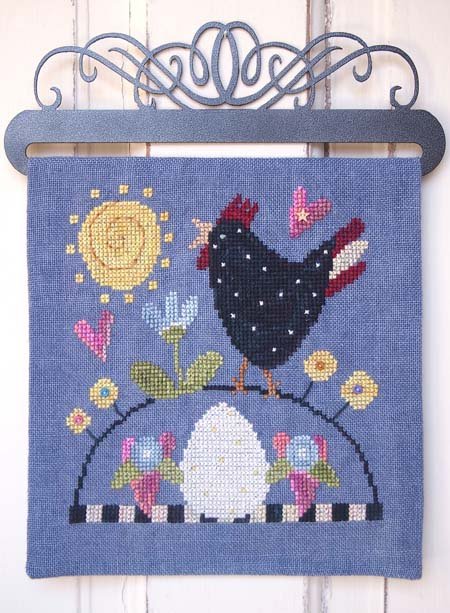 A square blue cross stitch featuring a cross-stitch style design of a black hen, white egg, yellow sun, pink and blue flowers, and small birds, hung from a dark metal hanger against a white wood plank wall.
Photo by SamSarah Studio Designs [image_link]