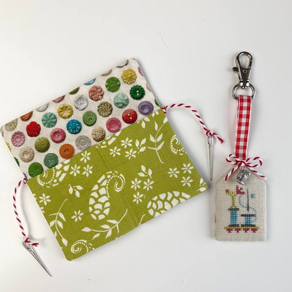 A handmade fabric needle case with a green and white floral pattern, decorated with colorful buttons on the flap, shown next to a matching embroidered cross-stitch sampler and a red-and-white gingham ribbon keychain on a white background.
Photo by HandsOnDesign.biz [additional_image_link]