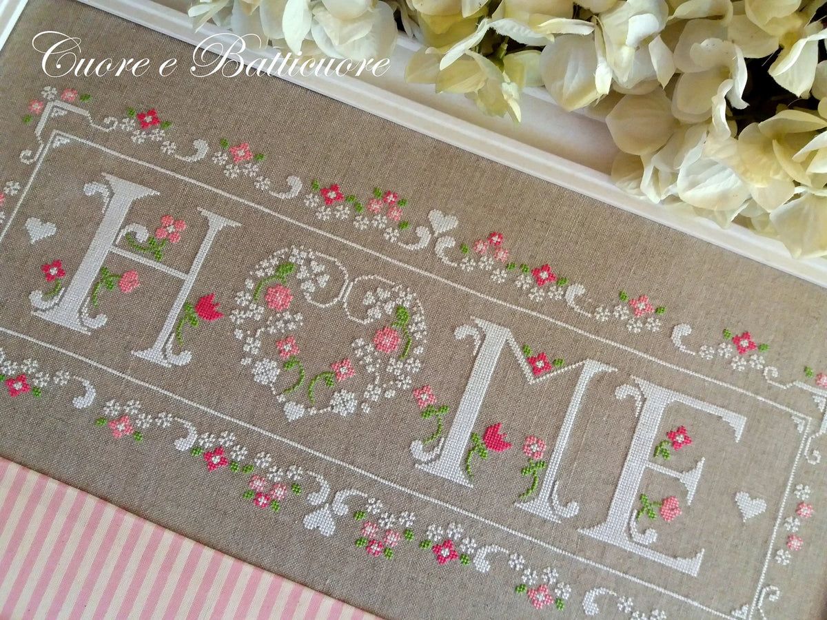 A close-up of a decorative embroidered textile with the word “HOME” in large white letters, surrounded by pink and white floral embroidery on a beige fabric background. The words “Enorme e Baltimore” appear in cursive at the top, and cream flowers are arranged along the upper edge.
Photo by CuoreBatticuore.com [additional_image_link]