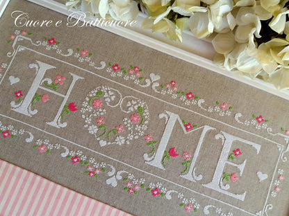 A close-up of a decorative embroidered textile with the word “HOME” in large white letters, surrounded by pink and white floral embroidery on a beige fabric background. The words “Enorme e Baltimore” appear in cursive at the top, and cream flowers are arranged along the upper edge.
Photo by CuoreBatticuore.com [additional_image_link]