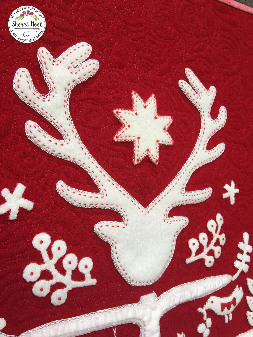 A close-up of a red quilted fabric featuring a white applique reindeer head with antlers, a white star, and white snowflake motifs, with a circular 'Sherri Noel' brand logo in the top-left corner.
Photo by Rebecca Mae Designs [additional_image_link]