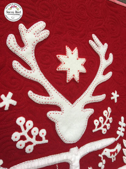 A close-up of a red quilted fabric featuring a white applique reindeer head with antlers, a white star, and white snowflake motifs, with a circular 'Sherri Noel' brand logo in the top-left corner.
Photo by Rebecca Mae Designs [additional_image_link]