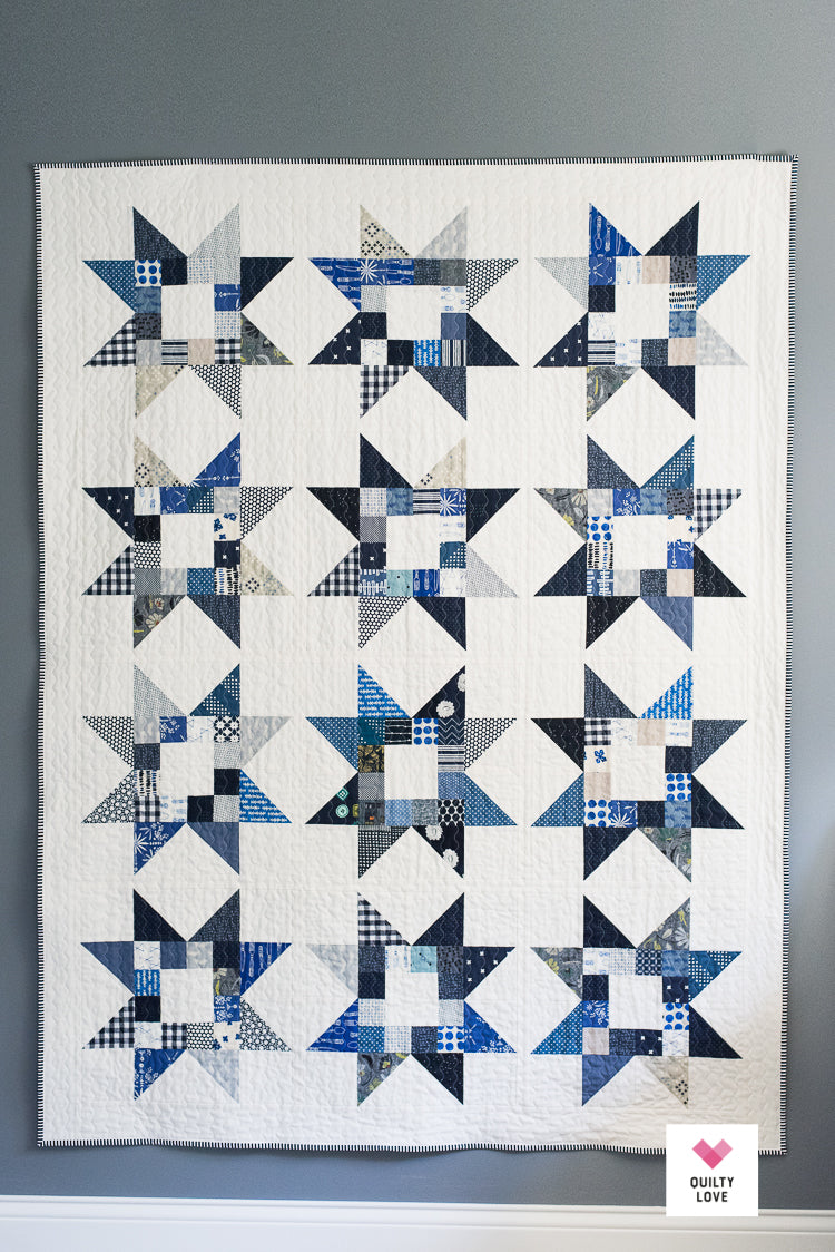 A handmade patchwork quilt featuring a repeating grid of blue and navy starburst patterns on an off-white background, hung on a gray wall.
Photo by Quilty Love [additional_image_link]