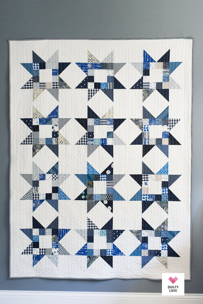 A handmade patchwork quilt featuring a repeating grid of blue and navy starburst patterns on an off-white background, hung on a gray wall.
Photo by Quilty Love [additional_image_link]