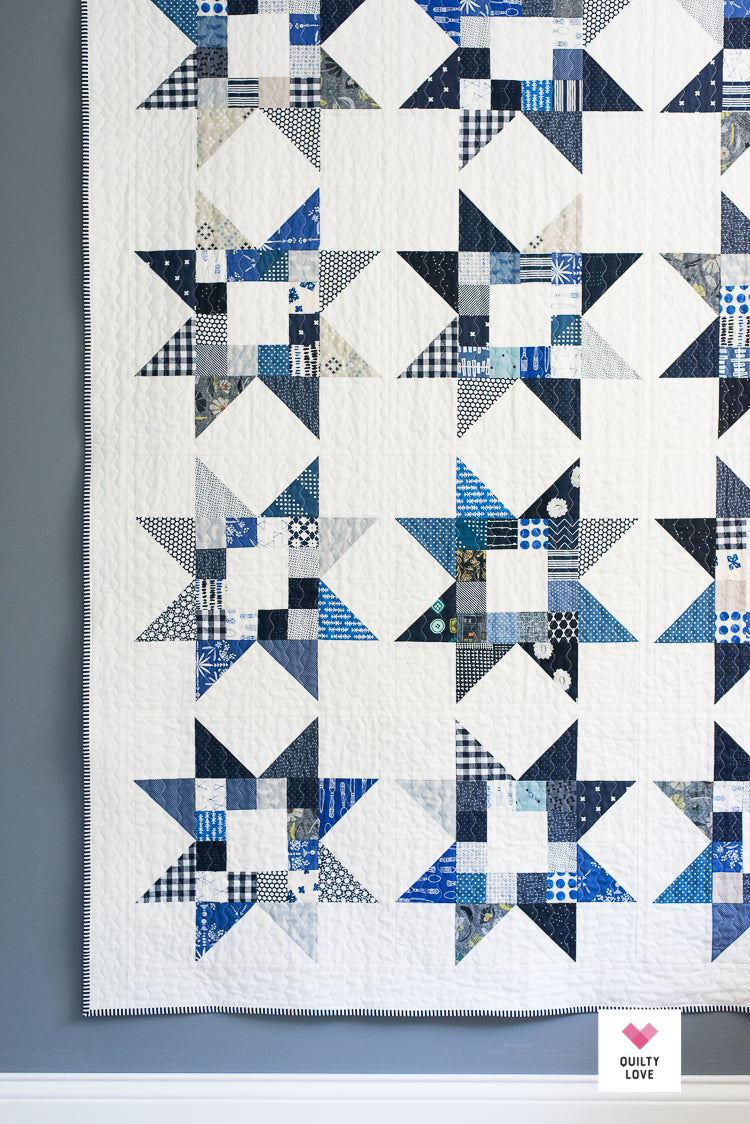 A quilted fabric panel featuring a repeating geometric starburst pattern in shades of blue, navy, gray, and white, with a visible 'Dusty Love' brand label in the bottom right corner.
Photo by Quilty Love [additional_image_link]