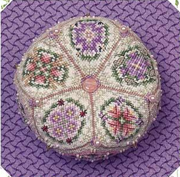 A round, hand-embroidered biscornu featuring six floral panels in purple, pink, green, and white on a cream background, with a central pink circle, displayed on a purple textured background.
Photo by JustNan.com [additional_image_link]