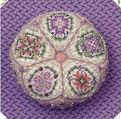 A round, hand-embroidered biscornu featuring six floral panels in purple, pink, green, and white on a cream background, with a central pink circle, displayed on a purple textured background.
Photo by JustNan.com [additional_image_link]