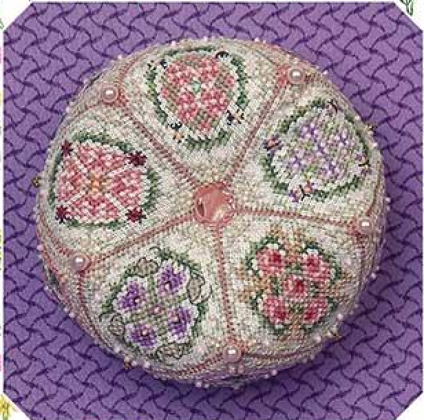 A round, hand-embroidered biscornu with floral patterns in pink, purple, and green on a cream background, decorated with small pearls and a pink bead at the center, set against a textured purple background.
Photo by JustNan.com [additional_image_link]