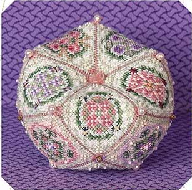 A vintage-style embroidered beaded biscornu with a floral pattern in pink, green, and purple on a white base, displayed on a textured purple background.
Photo by JustNan.com [additional_image_link]