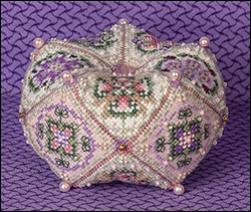 A hand-beaded, biscornu with intricate floral patterns in pink, green, and purple on a white base, set against a textured purple background.
Photo by JustNan.com [additional_image_link]