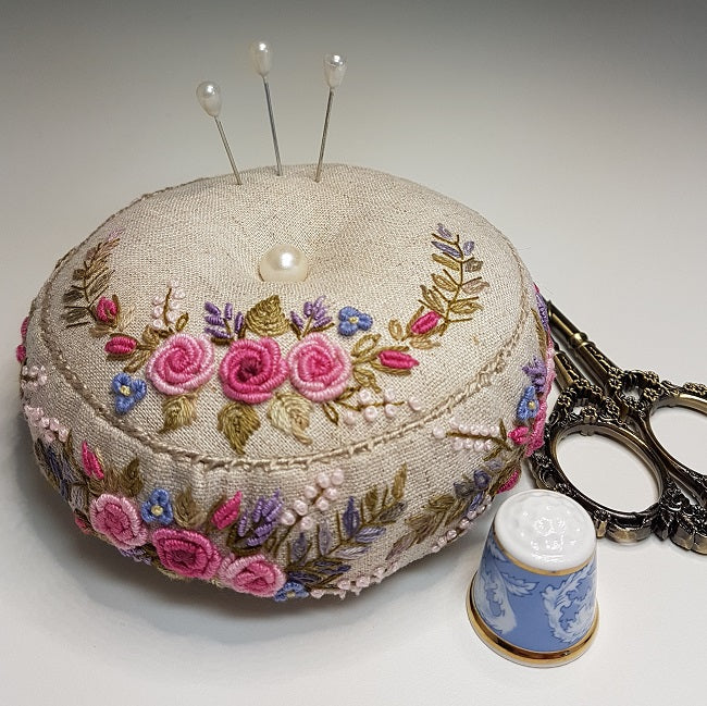 An embroidered round pincushion made of beige linen fabric, decorated with pink and purple roses, blue flowers, and green leaves. It is shown with three silver pins, ornate silver scissors, and a blue-and-white ceramic thimble on a light gray background.
Photo by Angela Watson [image_link]
