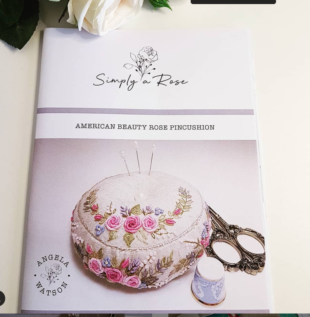 A book titled 'Simply a Rose: American Beauty Rose Pincushion' by Angela Watson, featuring a cover photo of a floral embroidered pincushion with pins, scissors, and a thimble on a light surface.
Photo by Angela Watson [additional_image_link]