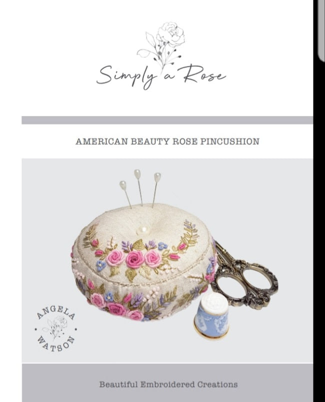 A product cover for 'American Beauty Rose Pincushion' by Simply a Rose, featuring an embroidered cream pincushion with pink and purple roses, decorative pins, ornate scissors, and a blue-and-white thimble on a light gray background.
Photo by Angela Watson [additional_image_link]