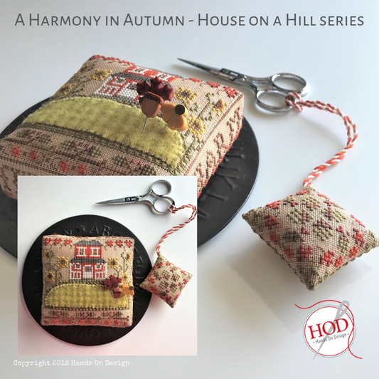 A handmade rectangular pincushion pillow with a floral and embroidered house design, shown alongside a matching small pincushion with a salmon-and-white cord. Scissors and a black plate are styling props on a white background.
Photo by HandsOnDesign.biz [image_link]