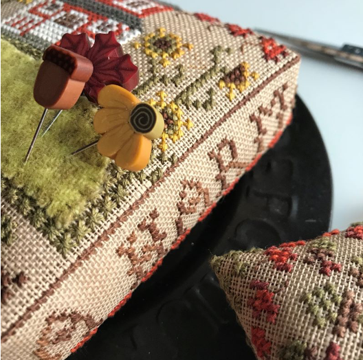 A close-up of a decorative cross-stitch pincushion with embroidered floral and letter motifs, topped with wooden flower-shaped pins.
Photo by HandsOnDesign.biz [additional_image_link]