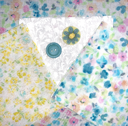 A close-up flat lay of several patterned fabric swatches with floral prints in pastel blue, yellow, pink, and green, plus two decorative buttons on top.
Photo by JustAnotherButtonCompany.com [additional_image_link]