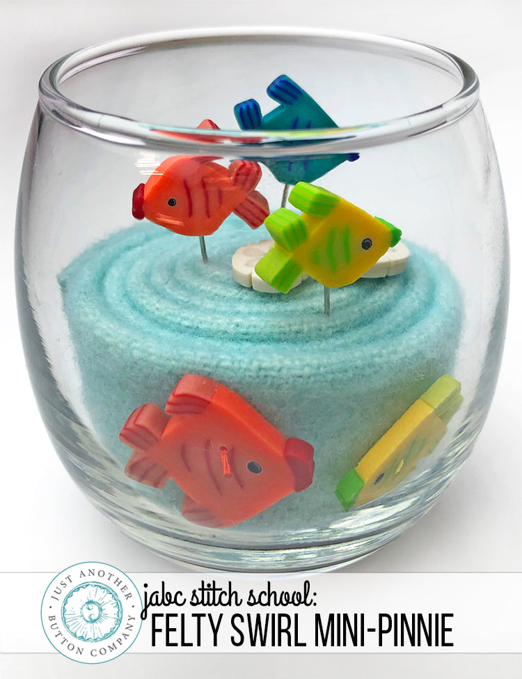 A clear glass bowl filled with light blue felt fabric and small colorful fish-shaped pins, shown on a white background with a 'jabc stitch school' logo at the bottom.
Photo by JustAnotherButtonCompany.com [image_link]