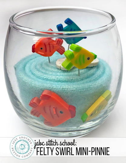 A clear glass bowl filled with light blue felt fabric and small colorful fish-shaped pins, shown on a white background with a 'jabc stitch school' logo at the bottom.
Photo by JustAnotherButtonCompany.com [image_link]