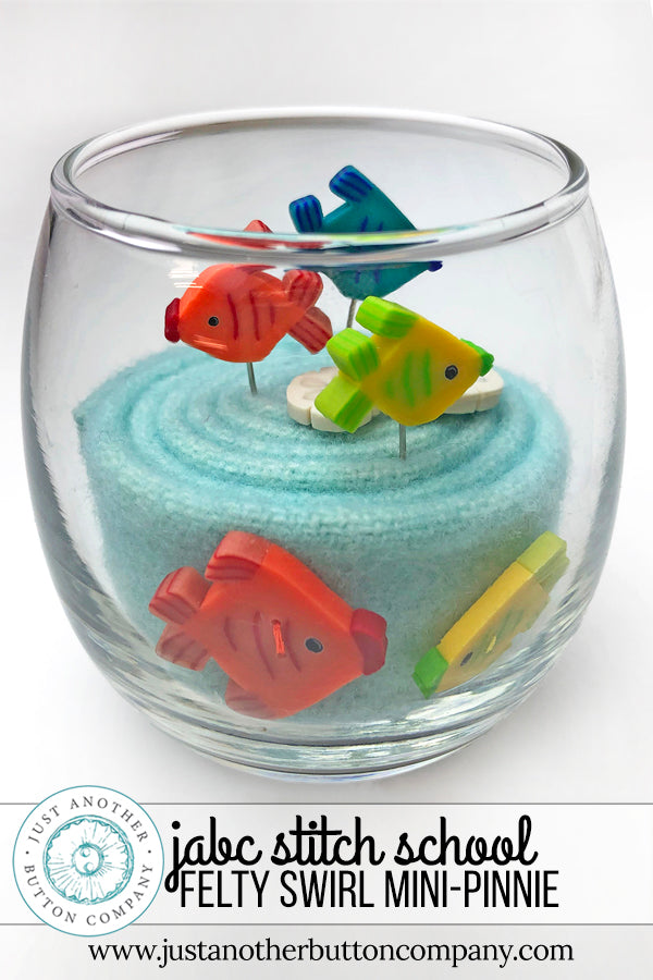 A clear glass bowl filled with light blue felt fabric and decorated with colorful felt fish pins, shown on a white background.
Photo by JustAnotherButtonCompany.com [additional_image_link]