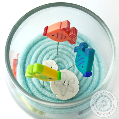 A top-down view of a glass bowl containing a spiral of light blue sand, with four colorful fish-shaped pins in orange, blue, green, and white attached to the rim.
Photo by JustAnotherButtonCompany.com [additional_image_link]