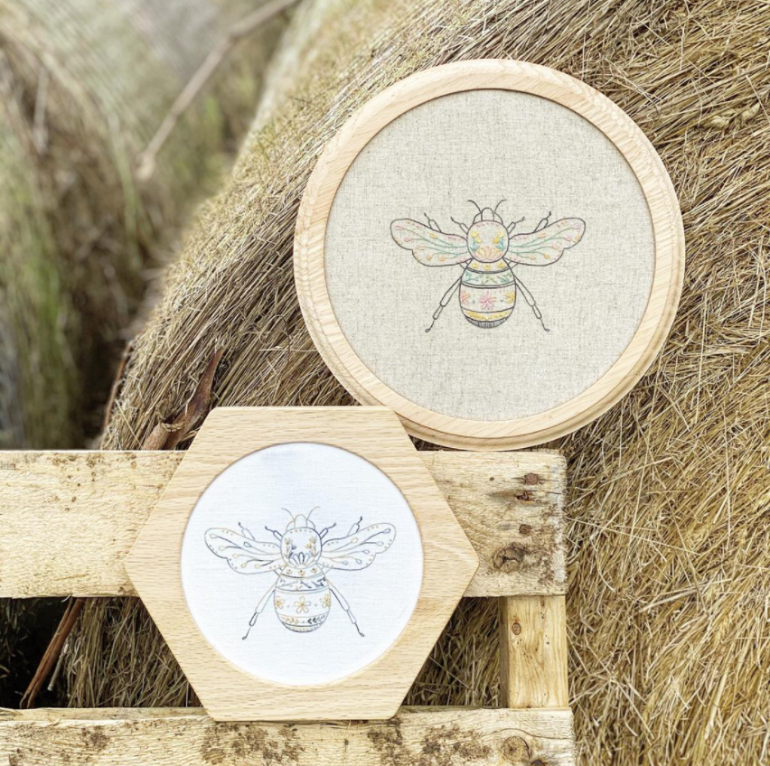 Two decorative wooden embroidery hoops with bumblebee designs, one round and one hexagonal, displayed on a rustic wooden bench in front of a hay bale.
Photo by Amy Kallissa [additional_image_link]