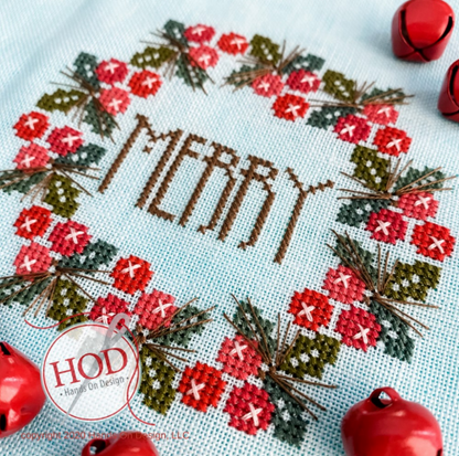 A close-up of a completed cross-stitch design featuring the word “MERRY” in brown thread, surrounded by a wreath of red flowers, green leaves, and brown pinecones on a light blue fabric. Red jingle bells are placed around the edges, and a circular watermark reads “HOD - Hand On Design.
Photo by HandsOnDesign.biz [image_link]