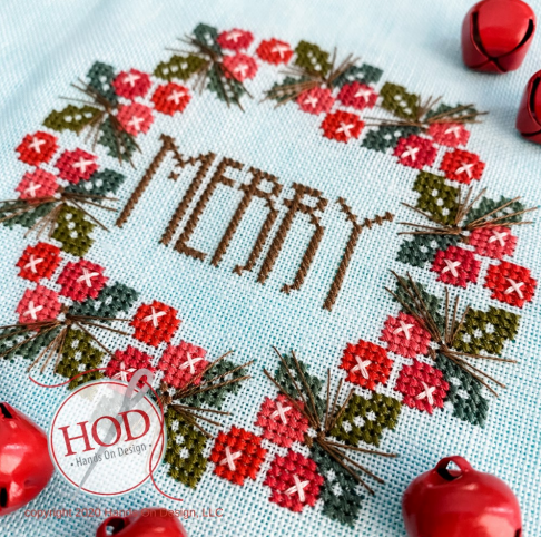 A close-up of a completed cross-stitch design featuring the word “MERRY” in brown thread, surrounded by a wreath of red flowers, green leaves, and brown pinecones on a light blue fabric. Red jingle bells are placed around the edges, and a circular watermark reads “HOD - Hand On Design.
Photo by HandsOnDesign.biz [image_link]