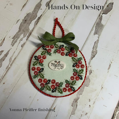 A round cross-stitch ornament featuring a floral wreath pattern in red and green on a white fabric background, with a small wooden 'Merry' button in the center and a green ribbon bow at the top. The ornament is displayed on a distressed white wooden surface with text overlays reading 'Hands On Design' and 'Vonna Pfeiffer finishing'.
Photo by HandsOnDesign.biz [additional_image_link]