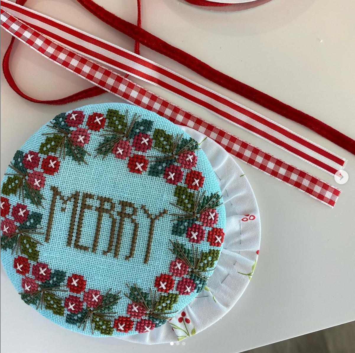 A round cross-stitch embroidery sample on light blue fabric with a red and green floral border and the word “MERRY” stitched in brown, shown on a white surface with red-and-white ribbon strips nearby.
Photo by HandsOnDesign.biz [additional_image_link]