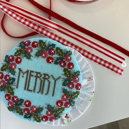 A round cross-stitch embroidery sample on light blue fabric with a red and green floral border and the word “MERRY” stitched in brown, shown on a white surface with red-and-white ribbon strips nearby.
Photo by HandsOnDesign.biz [additional_image_link]
