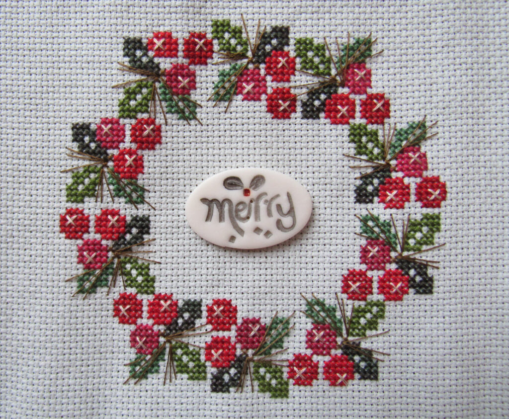 A handmade embroidered Christmas wreath on white Aida, featuring red berries, green and dark gray pine needles, and a central white oval button with 'merry' stitched in black.
Photo by HandsOnDesign.biz [additional_image_link]