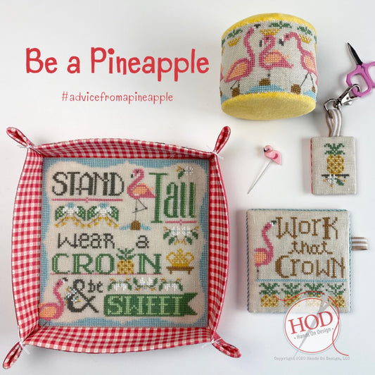 A coordinated set of cross-stitch themed items on a white background, including a framed square cushion with red gingham trim and pineapple/flamingo embroidery, a matching cylindrical pouch, a small square pouch, a pin, and a keychain. Text reads 'Be a Pineapple #advicefromapineapple' and a logo at bottom right says HOD craft by design.
Photo by HandsOnDesign.biz [image_link]