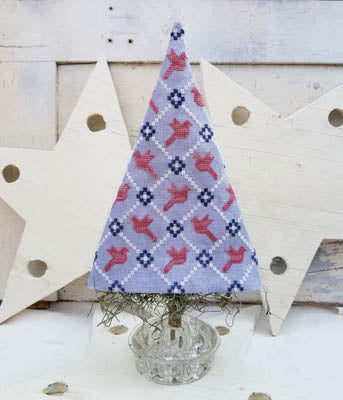 Cross stitch kit depicting a Christmas tree with red and blue checkered patterns on a white background, displayed on a small tree stand.
Photo by HelloFromLizMathews.com [image_link]